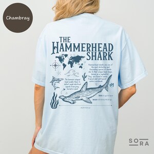K&ouml;nnte beinhalten: Hellblaues T-Shirt mit einem Grafikdruck eines Hammerhais und dem Text "The Hammerhead Shark". Die Grafik zeigt eine Weltkarte, Wellen und einen Hammerhai mit dem Text "Speed: 25 mph", "Weight: 200-300 lbs (90-135 kg)" und "0-20 ft (0-6.4 m)".