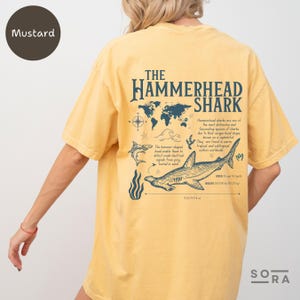 K&ouml;nnte beinhalten: Ein senfgelbes T-Shirt mit einem Grafikdruck eines Hammerhais und dem Text "THE HAMMERHEAD SHARK". Die Grafik enth&auml;lt eine Weltkarte, einen Kompass und einen Hammerhai mit Informationen &uuml;ber die Art.