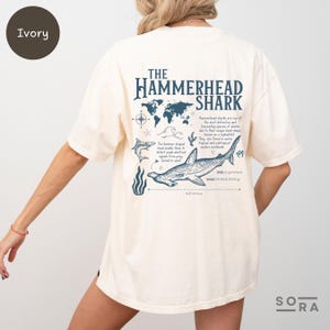 K&ouml;nnte beinhalten: Ein T-Shirt in Elfenbein mit einer blauen Grafik eines Hammerhais und dem Text "The Hammerhead Shark". Die Grafik enth&auml;lt auch eine Weltkarte, einen Kompass und eine Beschreibung des Hammerhais.