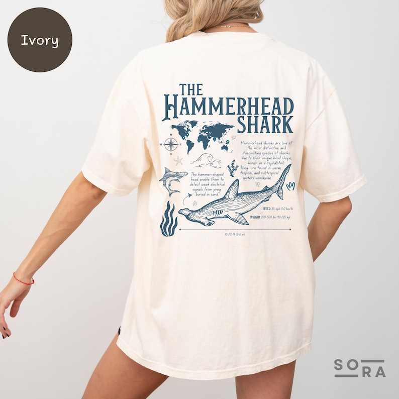 K&ouml;nnte beinhalten: Ein T-Shirt in Elfenbein mit einer blauen Grafik eines Hammerhais und dem Text "The Hammerhead Shark". Die Grafik enth&auml;lt auch eine Weltkarte, einen Kompass und eine Beschreibung des Hammerhais.