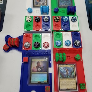 May include: A blue and red 3D printed game organizer with dice, cards, and a command zone. The cards are "The Earthshaker Kiora" and "Alistair, the Brigadier".