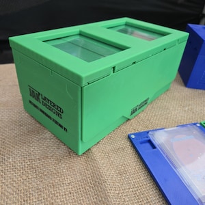 May include: A bright green storage box with two transparent windows on top. The box has a rectangular shape and features the text "LAYERED DESIGNS" and "AMG GAME CARD STATION V2". Other blue and green boxes are also visible.