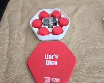 May include: A red and white Liar's Dice game set. The set includes a red hexagonal lid with the game title in white, a white container with red dice cups, and dice. The game is produced by Layered Designs.