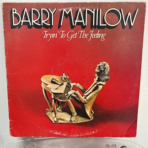 May include: A vinyl record album cover featuring Barry Manilow. The cover is red with a gold-colored sculpture of a person sitting at a table. The album title is "Tryin' To Get The Feeling".