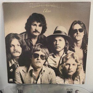 May include: A vintage vinyl record album cover featuring a band of seven men. The album title "Firefall" and the word "Elan" are visible. The men are dressed in 1970s attire, with long hair and sunglasses.