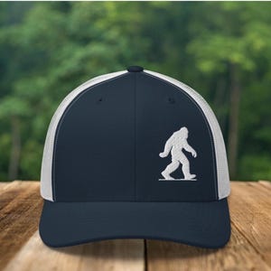 Könnte beinhalten: Marineblaue Trucker-Kappe mit weißem Mesh-Rücken und einer weißen, gestickten Bigfoot-Silhouette. Die Kappe hat einen gebogenen Schirm und einen Snapback-Verschluss. Die Kappe liegt auf einer Holzoberfläche.