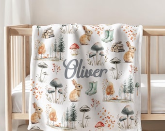 May include: A white blanket featuring a repeating pattern of brown rabbits, various mushrooms, green rain boots, stacks of logs, and forest trees. The name Oliver is printed in a gray script font in the center of the blanket.
