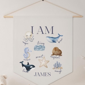 May include: A cream-colored wall hanging with a gold rod, featuring the words "I AM" in navy blue. Below are watercolor illustrations of sea creatures and words like "unique" and "brave". The name "JAMES" is printed at the bottom.