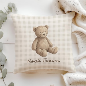 Peut inclure: Coussin carré avec un motif vichy beige clair et une illustration à l'aquarelle d'un ours en peluche. L'ours est brun et assis. Le nom "Noah James" est imprimé en gris foncé.