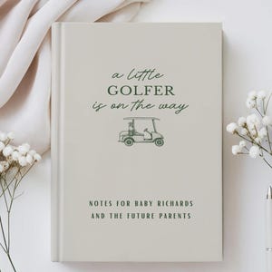 May include: A light gray notebook with the text "a little GOLFER is on the way" and a golf cart illustration in green. Below the text is "NOTES FOR BABY RICHARDS AND THE FUTURE PARENTS". The notebook is surrounded by white flowers, a pen, and a cup of coffee.
