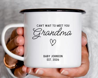Puede incluir: Taza de esmalte blanco con borde y asa negros. La taza tiene el texto "CAN'T WAIT TO MEET YOU Grandma" con un símbolo de corazón y "BABY JOHNSON EST. 2026" impreso en negro. La taza está sostenida en las manos de una persona.