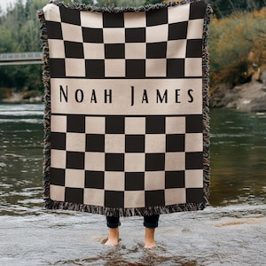 May include: A black and beige checkered throw blanket with the name "NOAH JAMES" in a beige band across the center. The blanket has a fringed edge and is held up by a person standing in a river.
