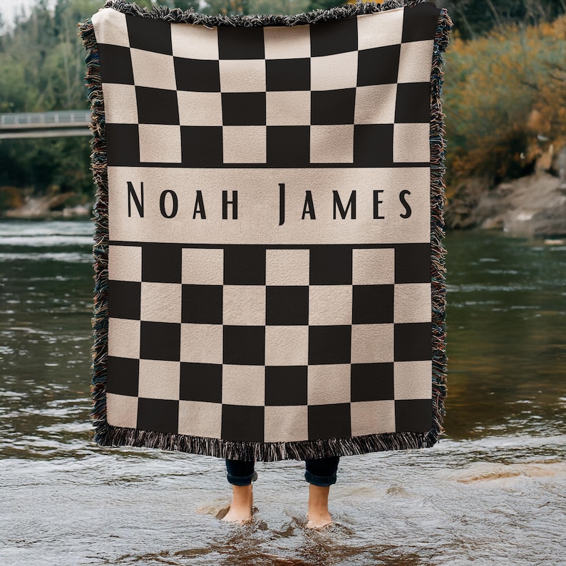 May include: A black and beige checkered throw blanket with the name "NOAH JAMES" in a beige band across the center. The blanket has a fringed edge and is held up by a person standing in a river.