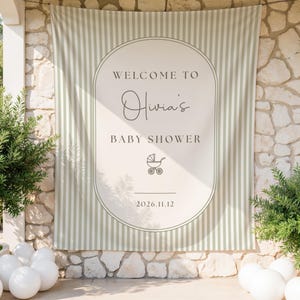 May include: A rectangular fabric backdrop featuring a sage green and white vertical striped pattern. The centre has a large cream-coloured oval frame with the text "WELCOME TO Olivia's BABY SHOWER", a small line-art baby carriage icon, and "2026.11.12".