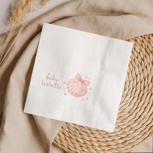May include: A square white paper napkin featuring a printed illustration of a light pink seashell adorned with a matching pink ribbon bow and small white pearls. The text "baby Isabella" is printed in a pink script font on the left side.