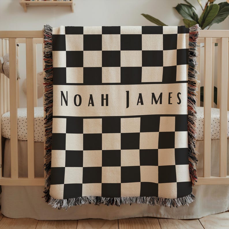 May include: A black and white checkered baby blanket with the name "NOAH JAMES" in black block letters. The blanket has a cream-colored background and a fringe border. The blanket is draped over a wooden crib.