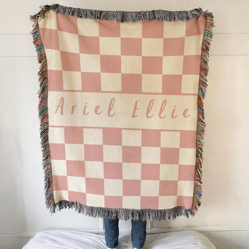 May include: A pink and cream chequered throw blanket with fringed edges. The blanket has the names "Ariel Ellie" in a cursive font across the centre. The blanket is displayed over a bed.