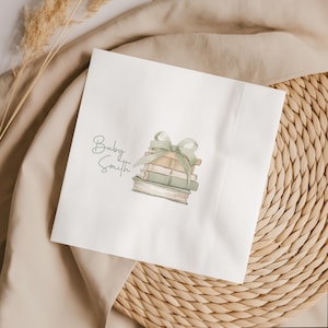 May include: A square white paper napkin featuring a watercolor illustration of a stack of three books tied with a sage green ribbon bow. The text "Baby Smith" is printed in a dark gray cursive script to the left of the book stack illustration.