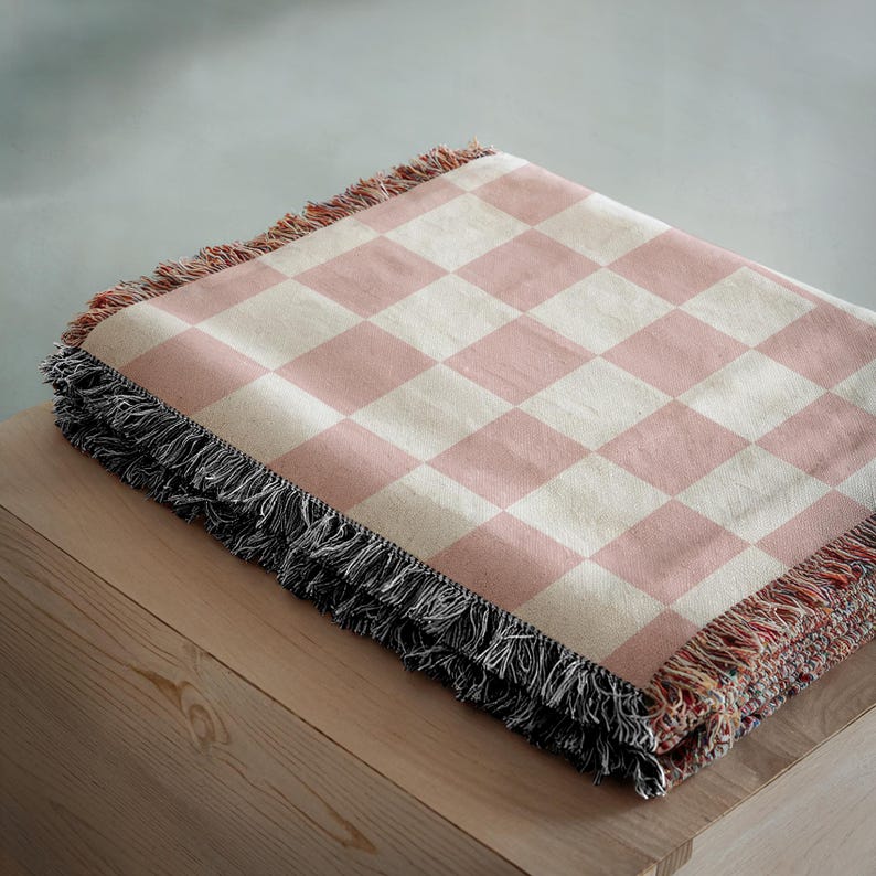 May include: A folded pink and cream-coloured checked throw blanket with a black fringe. The blanket is made of a soft material and is folded neatly on a wooden surface. The blanket's pattern is a classic checkerboard design.