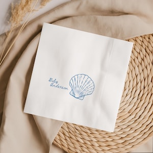 May include: A square white paper napkin featuring a blue line-art illustration of a seashell and the text Baby Anderson printed in a cursive script font. The napkin has a textured border and a smooth center area where the design is placed.