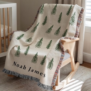 Puede incluir: Una manta crema con un patrón repetido de árboles verdes está sobre una mecedora de madera. La manta tiene un borde con flecos multicolores y el nombre "Noah James" está impreso en negro.