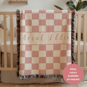 May include: A pink and cream checkered baby blanket with the names "Ariel Ellie" printed across the centre. The blanket has a fringed edge and is draped over a wooden cot. The text "Other colours available on request" is in the bottom right corner.