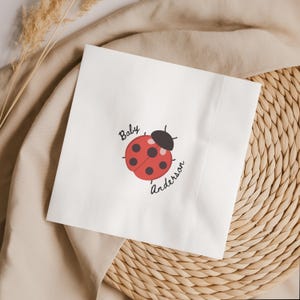 May include: White square paper napkin with a red ladybug illustration and the text "Baby Anderson". The ladybug has black spots and a black head. The napkin is on a beige surface.