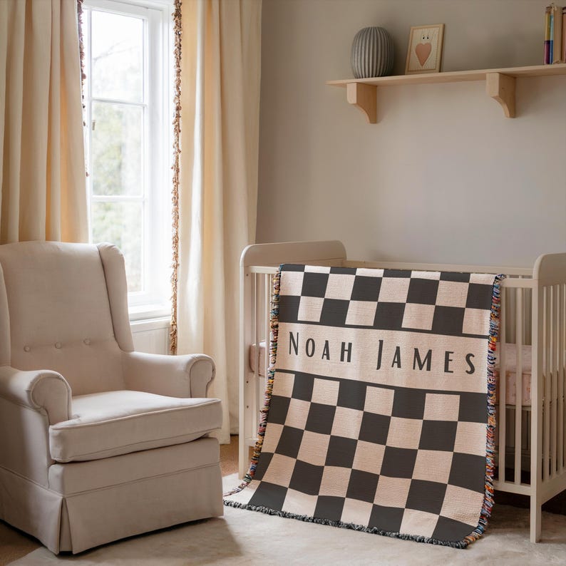 May include: A nursery scene with a cream-colored crib and a personalized blanket. The blanket features a black and cream checkerboard pattern with the name "NOAH JAMES" in bold letters. A beige armchair sits nearby.