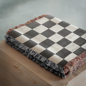 May include: A folded blanket with a black and cream checkerboard pattern. The blanket has a fringed edge with black, red, and cream threads. The blanket is folded on a wooden surface.
