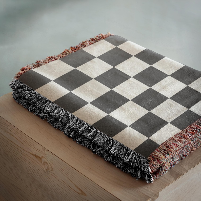 May include: A folded blanket with a black and cream checkerboard pattern. The blanket has a fringed edge with black, red, and cream threads. The blanket is folded on a wooden surface.