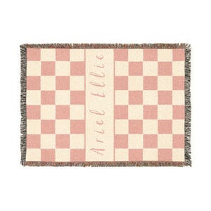 May include: A woven throw blanket featuring a pink and cream checkerboard design. The blanket has a fringed edge, and the name "Ariel Ellie" is printed vertically in a script font down the centre.