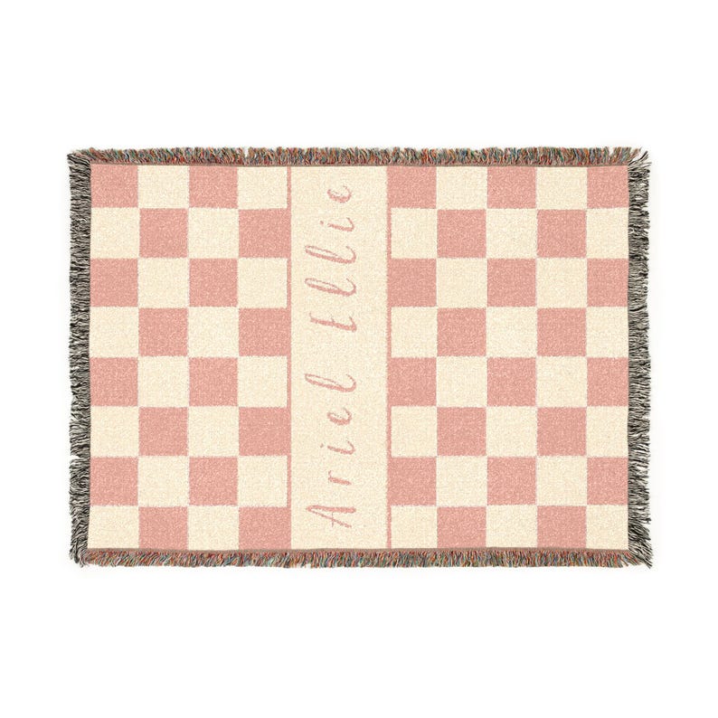 May include: A woven throw blanket featuring a pink and cream checkerboard design. The blanket has a fringed edge, and the name "Ariel Ellie" is printed vertically in a script font down the centre.