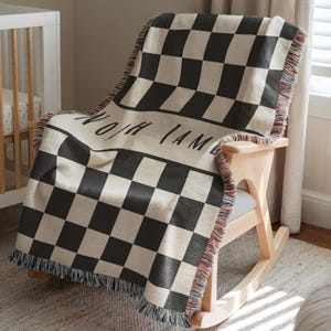 May include: A black and white checkered throw blanket with fringe is draped over a wooden rocking chair. The blanket has the name "NOAH I AM" printed on it. The blanket is made of a soft material.