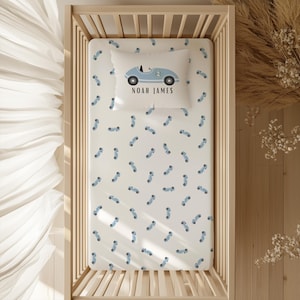 May include: A cot sheet and pillowcase set with a pattern of blue race cars on a white background. The pillowcase has a larger car design with the name "NOAH JAMES" and the number "2". The bedding is inside a wooden cot.