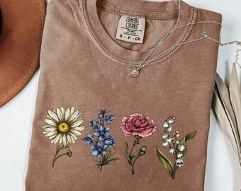 Op de afbeelding: Een bruin T-shirt met een bloemenprint met een madeliefje, een blauwe bloem, een roze anjer en lelietje-van-dalen. Het shirt heeft een label met de tekst "Comfort Colors" en "S-P-CH".