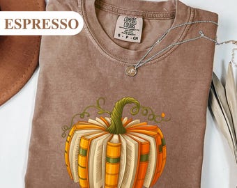 Könnte beinhalten: Espressofarbenes T-Shirt mit einem Kürbisdesign aus Büchern. Der Kürbis hat orange und gelbe Seiten mit grünen Ranken und einem grünen Stiel. Das T-Shirt hat einen Rundhalsausschnitt und kurze Ärmel. Eine silberne Halskette mit Anhänger ist ebenfalls sichtbar.