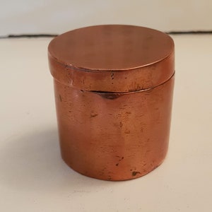 Peut inclure: Petit récipient cylindrique en cuivre avec couvercle assorti. Le récipient a une surface lisse et polie et une couleur brun-rougeâtre chaude. Le couvercle s'adapte parfaitement. Idéal pour ranger de petits objets. Hauteur d'environ 7 cm.