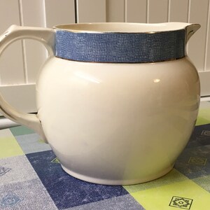 May include: Cream-colored ceramic pitcher with a rounded body and a curved handle. The top band is a textured blue with gold trim. The pitcher has a spout for easy pouring and is likely used for serving beverages.