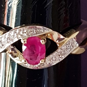 May include: A gold ring with a central oval ruby gemstone. The ring features a twisted design with a band of small, clear gemstones on either side of the ruby. The ring is displayed on a black stand.
