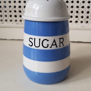 May include: A ceramic sugar shaker with a white lid and a blue and white striped body. The word "SUGAR" is printed in black on the front. The shaker is a kitchenware item.