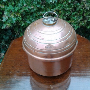 Puede incluir: Una olla de cocina de color cobre, de forma esférica, con un asa en la parte superior. La olla tiene estrías circulares concéntricas y la palabra "WAFAX" está estampada en el lateral. La olla está sobre una superficie de madera.