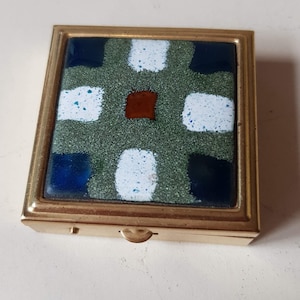 May include: A square, gold-coloured metal compact with a colourful enamel top. The enamel design features a green speckled background with blue, white, and dark blue squares, and a central brown square. The compact is closed.