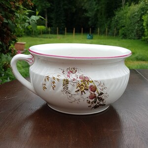 Puede incluir: Orinal antiguo blanco con asa y diseño floral. El borde está adornado con un borde rosa. El orinal está sobre una superficie de madera marrón oscuro, con un fondo verde.