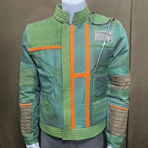 Peut inclure: Veste verte avec des détails orange et un col montant. La veste présente un motif texturé sur les épaules et les manches, avec des lignes horizontales et un emblème circulaire. Fermeture éclair et petite poche poitrine.