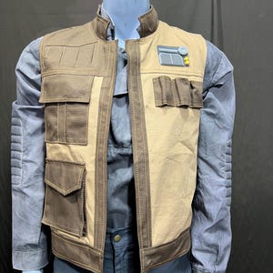 Peut inclure: Un gilet beige et marron avec plusieurs poches et un petit patch rectangulaire. Le gilet est porté sur une chemise grise avec un pantalon bleu foncé. Le gilet a un design militaire ou spatial.