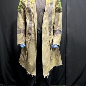 Może przedstawiać: Długi, beżowy płaszcz z frędzlami na dole. Płaszcz ma zamek błyskawiczny i kaptur. Płaszcz ma zielono-brązowe pasy na rękawach i plecach.