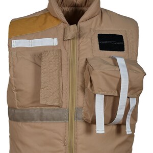 Peut inclure: Gilet matelassé de couleur beige avec plusieurs poches et un col montant. Il est doté d'une fermeture éclair intégrale, d'un empiècement d'épaule en daim marron et de sangles blanches.