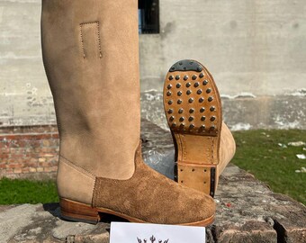 May include: A pair of brown leather boots with a suede toe and a studded sole. The boots are sitting on a brown stone surface. Rustic Clothing is printed on a white card in front of the boots.