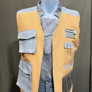 Peut inclure: Un gilet marron et bleu avec plusieurs poches et un col. Le gilet a un devant marron avec des poches et des accents bleus. Le gilet est présenté sur un mannequin. Le gilet a un design futuriste.
