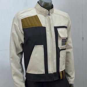 Peut inclure: Veste beige, noire et vert olive avec un col montant et une fermeture éclair. La veste présente un empiècement matelassé aux épaules, une poche poitrine et une ceinture marron et beige. La veste est présentée sur un mannequin.